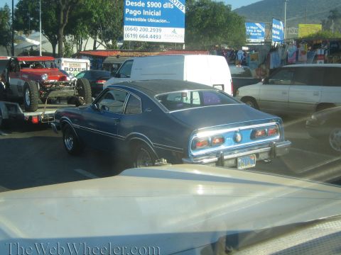 1990 Ford Bronco - dark blue 1970s two-door coupe sits in traffic with a red off-road buggy on a trailer at left and roadside vendor stalls behind.
