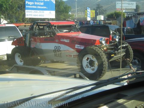 1990 Ford Bronco - red tube-frame off-road buggy with BFGoodrich and Old Guys Rule lettering rides on a car-hauler trailer amid roadside traffic.