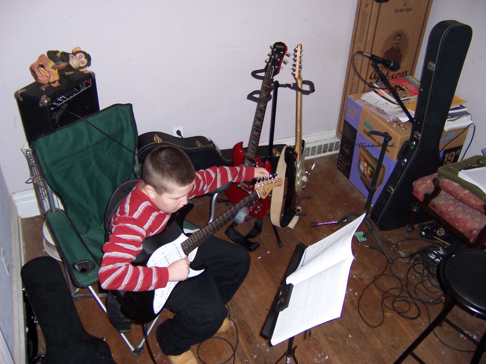 Boy in red and white striped shirt sits in a green chair, playing a white electric guitar. Several other guitars are on stands in the background