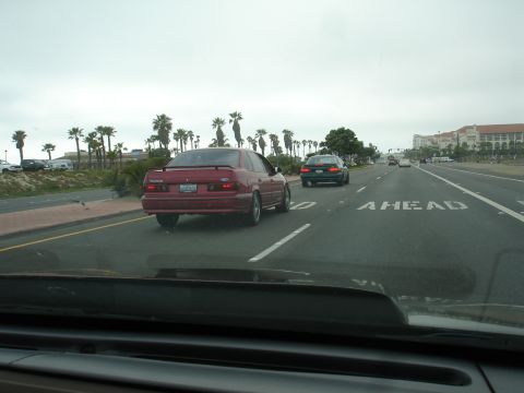 1993 Ford Taurus - A red sedan drives on a highway with palm trees and buildings in the background.