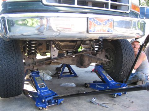 1974 Ford F-100 - A man works on the front suspension of a truck, which is elevated on jack stands and hydraulic jacks.