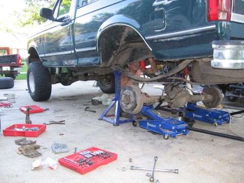 1974 Ford F-100 - A mechanic works on the rear suspension of a green Ford F-Series truck, which is raised on jack stands.