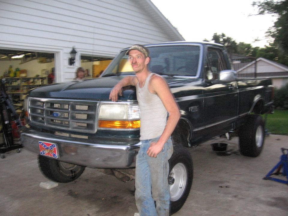 Guy leaning on a dark green Ford F-150 pickup truck. It has a lifted suspension and large tires