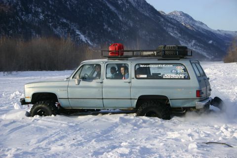 2003 Jeep TJ - A light blue SUV navigates through a snowy landscape, kicking up snow as it moves.