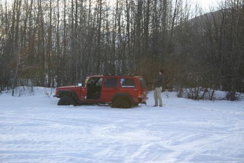 2003 Jeep TJ - A red Jeep Cherokee is partially buried in snow, with its driver's door ajar, as a person stands nearby in a snowy, wooded area.