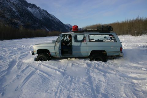 2003 Jeep TJ - An older model light blue SUV navigates through deep snow with its driver's door ajar, equipped with a roof rack and an auxiliary gas can.
