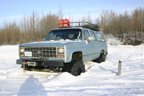 2003 Jeep TJ - Light blue Chevrolet Suburban with oversized tires and a roof rack with gas cans, stuck in snow.