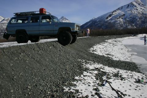 2003 Jeep TJ - A blue SUV with a roof rack and a red gas can is parked on a rocky shore with snow and mountains in the background.