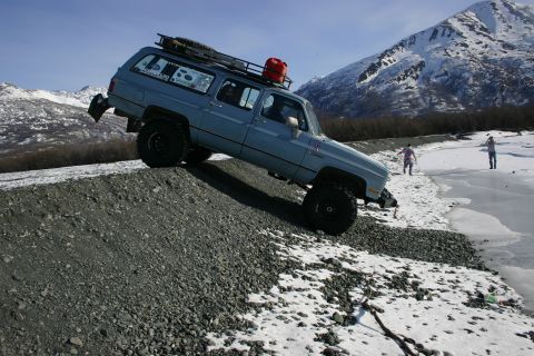 2003 Jeep TJ - A lifted blue SUV navigates a rocky incline near a partially frozen shoreline with snow-capped mountains in the distance.