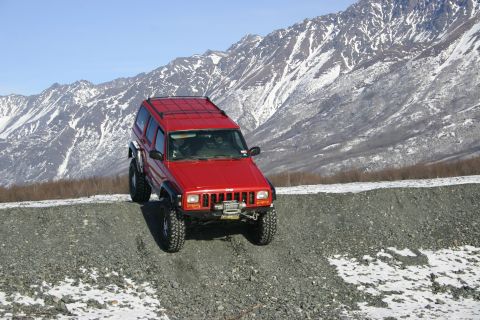 2003 Jeep TJ - A red Jeep Cherokee XJ navigates a gravel incline with a snow-dusted mountain range in the background.