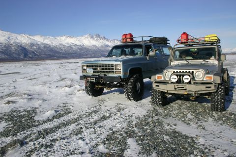 2003 Jeep TJ - Two modified off-road vehicles, a Jeep Wrangler and a Chevrolet Suburban, are parked in a snowy, mountainous area under a clear blue sky.