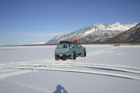 2003 Jeep TJ - SUV driving on a frozen lake with snow-covered mountains under a clear blue sky.