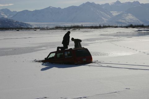 2003 Jeep TJ - Two people stand on the roof of a red SUV that is stuck in the ice, with snow-covered mountains in the background.