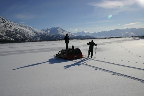 2003 Jeep TJ - Two people stand near a red SUV that is stuck in the snow with a mountain range in the background.