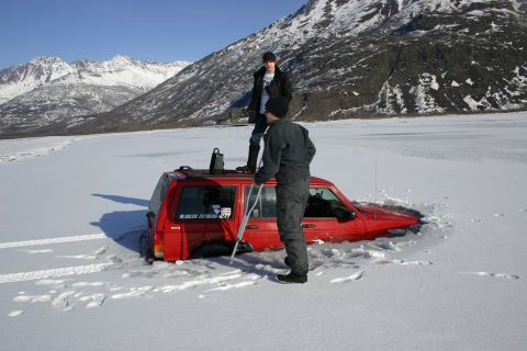 2003 Jeep TJ - A red SUV is partially submerged in ice, with two individuals standing nearby. One person is on the roof, and the other is beside the vehic…