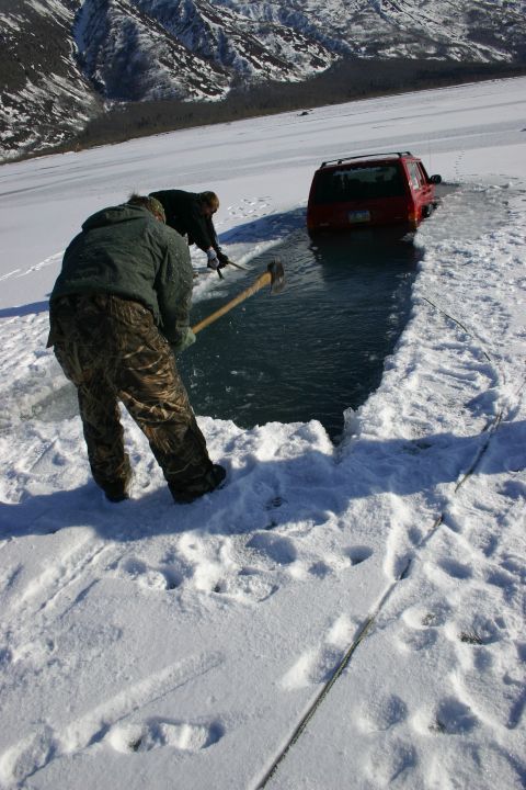 2003 Jeep TJ - Two individuals work to break ice around a red SUV that has become stuck in a frozen body of water.