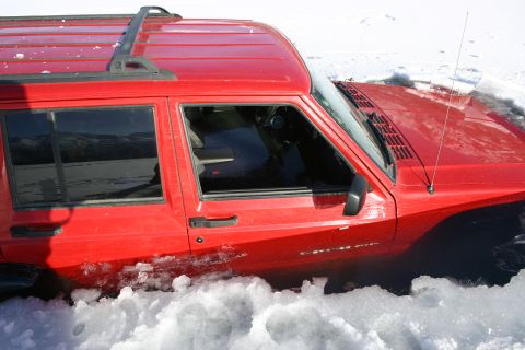 2003 Jeep TJ - A red Jeep Cherokee XJ is partially submerged in snow, showing its side and front quarter panel.