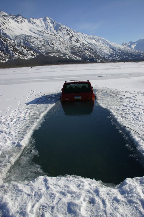 2003 Jeep TJ - A red SUV is partially submerged in a hole cut into the ice on a frozen body of water, with snow-capped mountains in the distance.