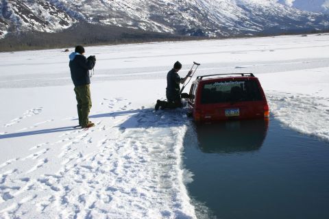 2003 Jeep TJ - A red SUV is partially submerged in icy water with two people nearby, one taking a photo and the other attempting to free the vehicle.