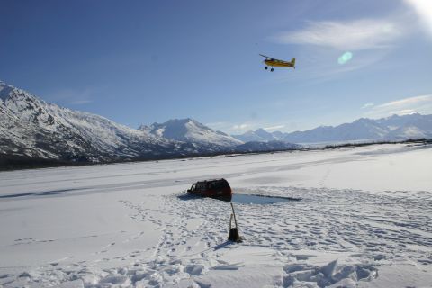 2003 Jeep TJ - A red SUV is partially submerged in a hole in the ice, with a small airplane flying overhead in a snowy mountainous region.