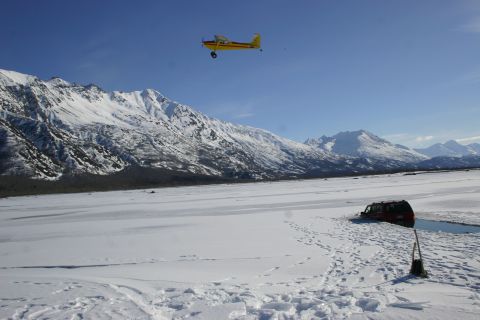 2003 Jeep TJ - A small plane flies over a vast, snow-covered landscape where a red SUV is partially submerged in icy water.