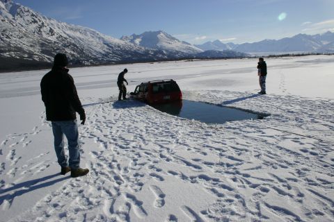 2003 Jeep TJ - A red 2003 Jeep TJ is partially submerged in an ice hole, with two people observing the scene.