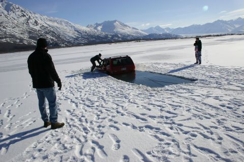 2003 Jeep TJ - A red SUV is partially submerged in a hole in the ice, with several people nearby appearing to attempt a rescue.
