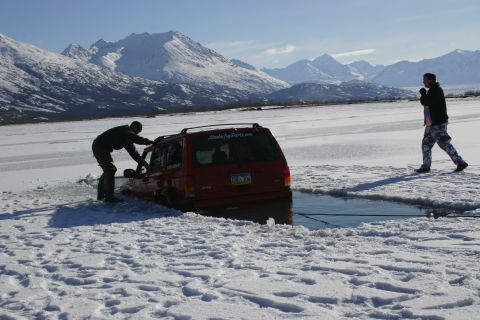 2003 Jeep TJ - A red SUV is partially submerged in icy water, with a person attempting to free it. Snow-covered mountains are visible in the distance.