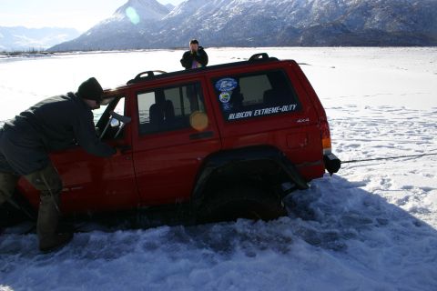 2003 Jeep TJ - A red Jeep Cherokee is stuck in deep snow near a frozen body of water, with two people nearby.