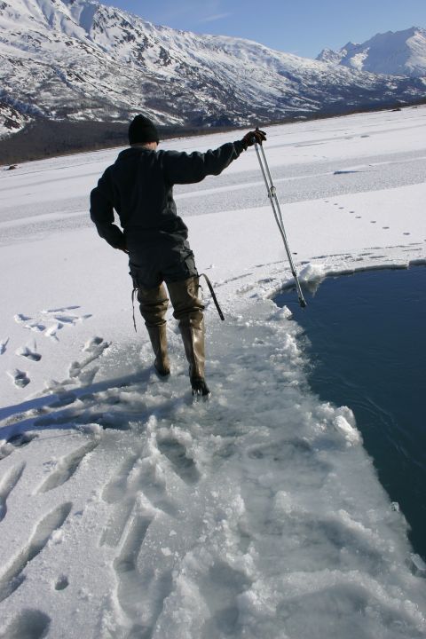2003 Jeep TJ - A person stands on an icy surface near open water, holding an ice pole, with snow-covered mountains in the distance.
