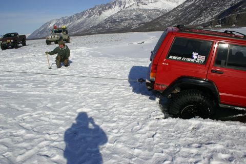 2003 Jeep TJ - A red Jeep Cherokee XJ is stuck in the snow while being winched by another vehicle, with mountains in the distance.