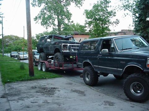 1992 Ford Bronco - A dark Ford Bronco tows another Bronco on a trailer, both appear modified with large tires.