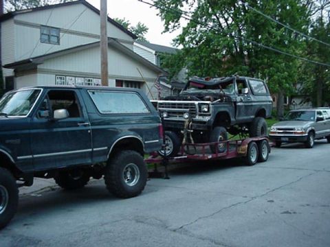 1992 Ford Bronco - A lifted dark teal 1990s Ford Bronco on chrome wheels and mud tires sits parked on a residential street towing a red tandem-axle flatbed tr…