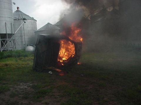 1992 Ford Bronco - A vehicle body shell lying on its side and fully engulfed in flames on a grassy farmyard, throwing heavy black smoke, with corrugated metal…