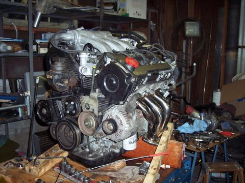1995 Ford Probe - Engine assembly with chrome exhaust headers sits on a wooden stand in a cluttered workshop environment.