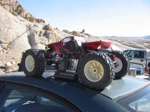 1985 Ford Bronco - A red remote control monster truck is positioned on top of a car, with a silver pickup truck visible in the background.