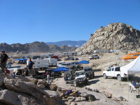 1985 Ford Bronco - A busy off-road gathering with various vehicles and campers set against a mountainous desert backdrop.