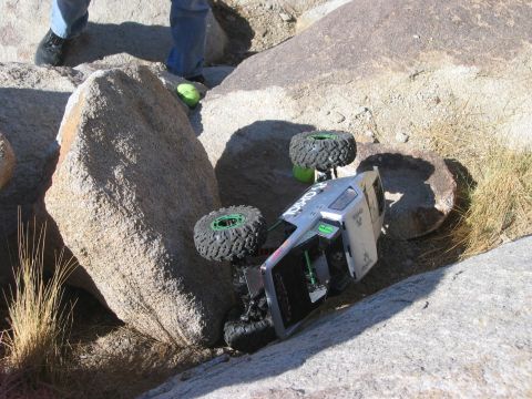 1985 Ford Bronco - A remote control rock crawler is stuck on its side amidst large boulders and dry grass.
