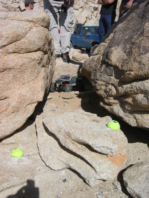 1985 Ford Bronco - A remote control rock crawler maneuvers through a narrow rocky passage, with a blue pickup truck visible in the background.