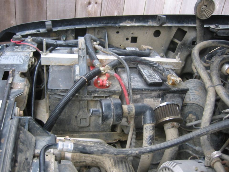 Dirty truck engine bay showing battery with red and black cables, hold-down bar and alternator
