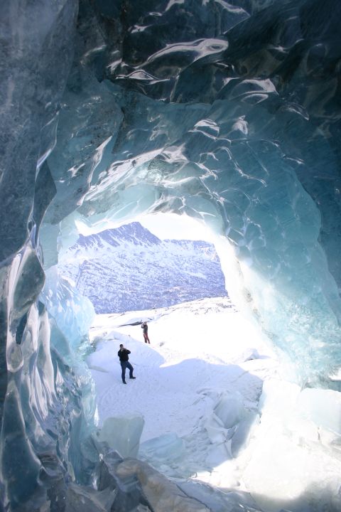2003 Jeep TJ - Two people explore the entrance of a glacial ice cave, with a vast snowy mountain landscape visible beyond.