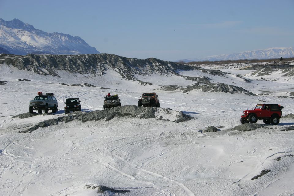 Five Jeep Wranglers drive across a snowy, rocky terrain with mountains in the background