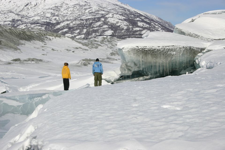 Two people stand on a snowy glacier, looking at ice formations and a mountain in the background