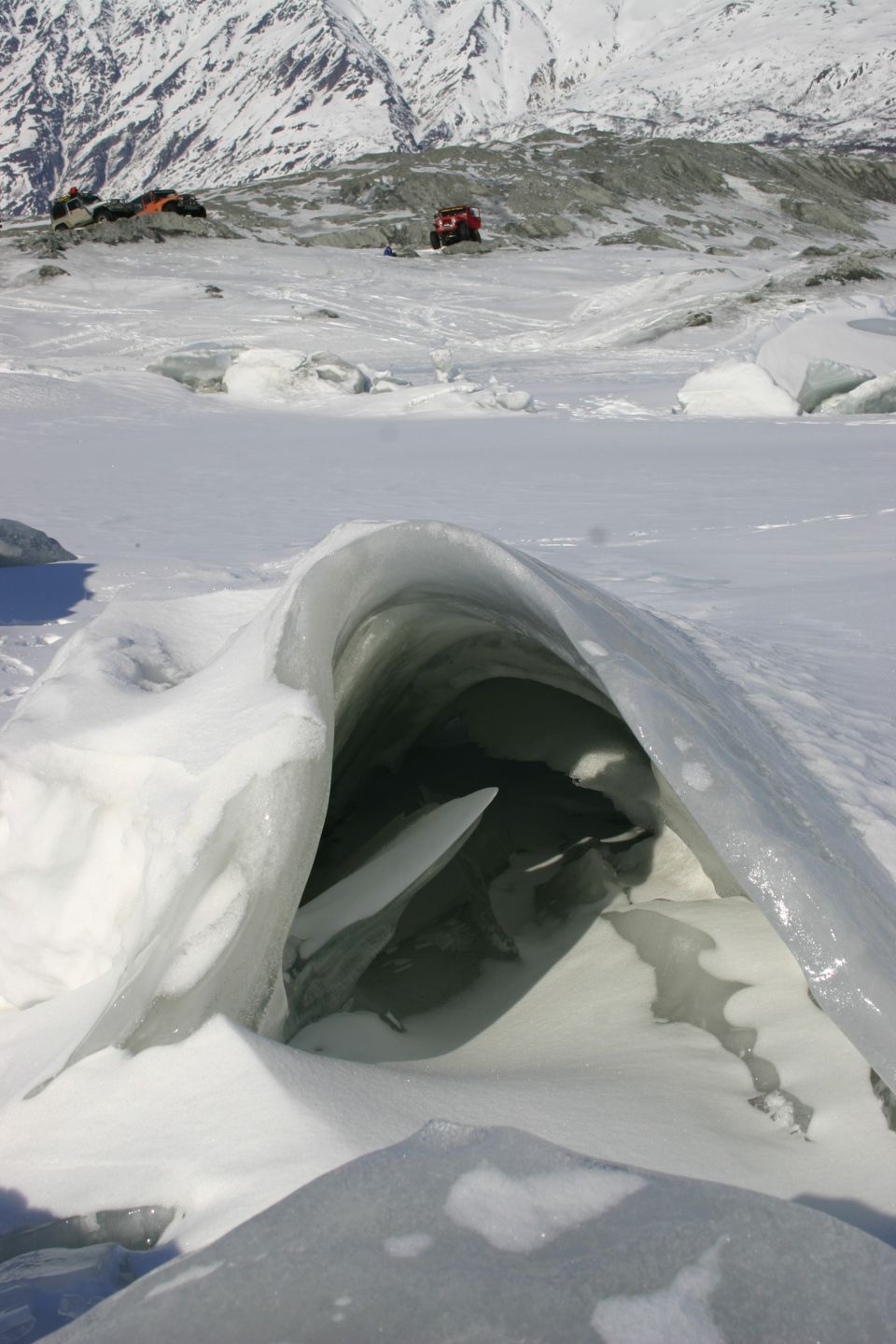 Ice cave entrance in a snowy landscape with mountains in the distance. Two vehicles and tents are visible on a ridge