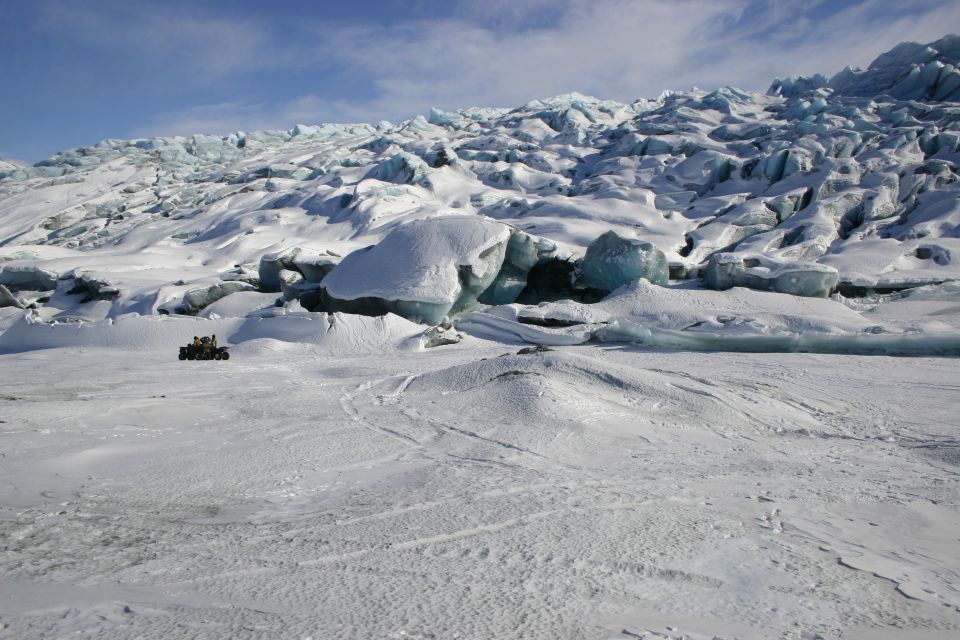 Snowmobile traversing a vast, icy glacier under a blue sky with scattered clouds