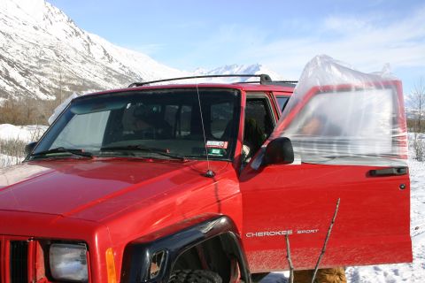 2003 Jeep TJ - A red Jeep Cherokee Sport sits in a snowy landscape with its driver's door open and covered in plastic sheeting.