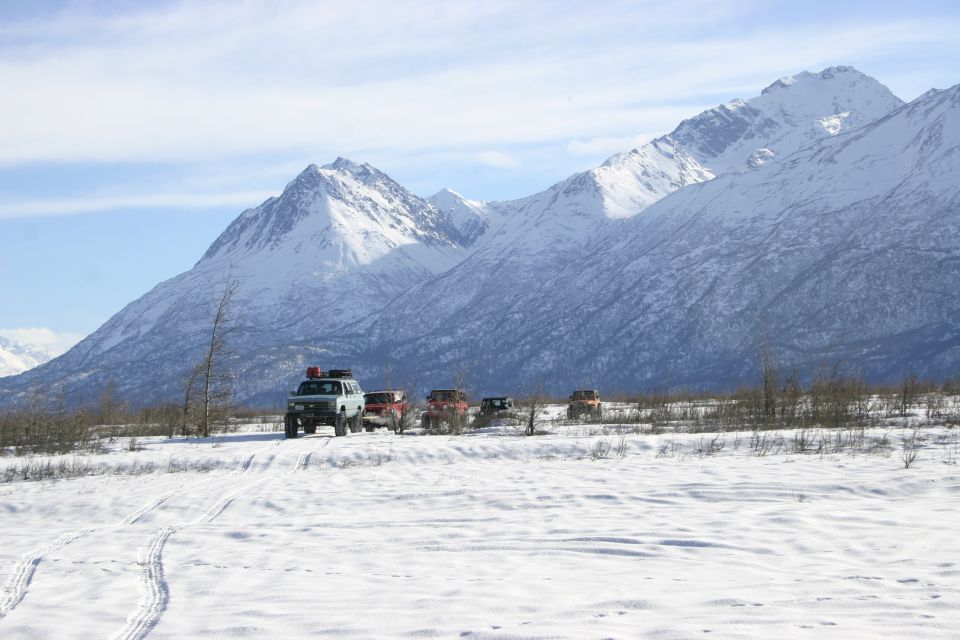 Several off-road vehicles drive through a snowy landscape with large, snow-covered mountains in the background