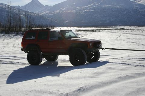 2003 Jeep TJ - A red Jeep Cherokee XJ, equipped with off-road tires, is being towed across a snowy landscape with mountains in the background.