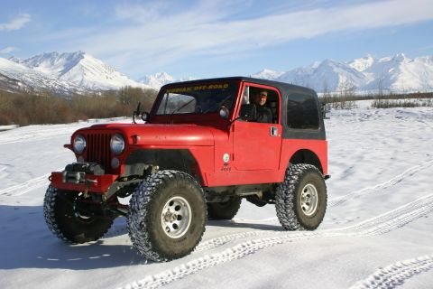 2003 Jeep TJ - Red Jeep CJ with black top and large tires on snow, mountains in background. "KODIAK OFF-ROAD" sticker on windshield.