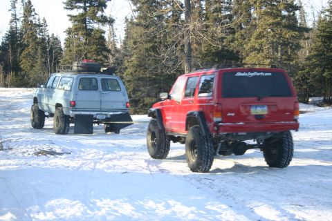 2003 Jeep TJ - A red Jeep Cherokee is being towed by a blue SUV through a snowy, wooded area.